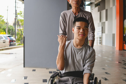 Young Asian man in a wheelchair pointing thumb back to guide direction while pushed by caregiver outdoors, concept of accessibility, communication, and independence.