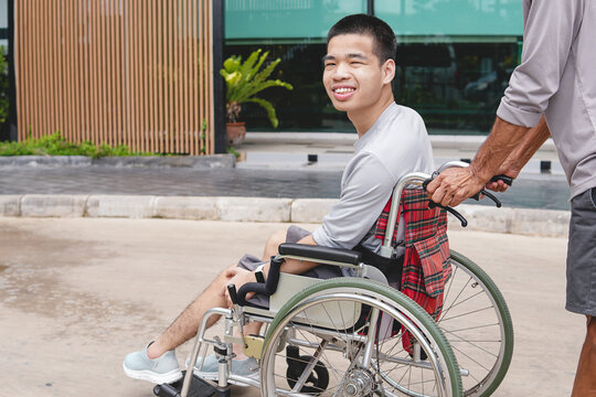 Happy young Asian man in a wheelchair looking at camera with a smile while pushed by caregiver outdoors, concept of positive attitude, accessibility, and inclusion.