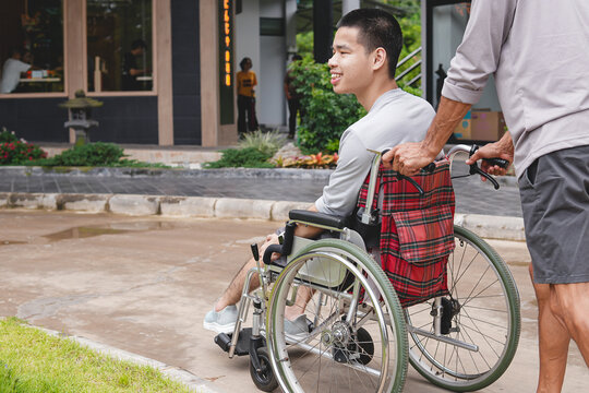 Happy young Asian man in a wheelchair pushed by a caregiver outdoors near a garden walkway, concept of accessibility, freedom, mobility, and inclusive lifestyle.