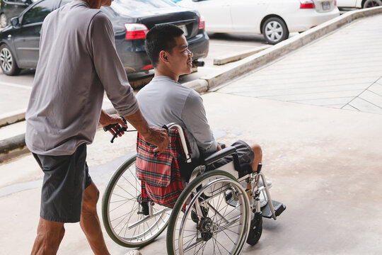 Asian man in wheelchair pushed by caregiver up ramp outdoors. Disabled son and father smiling together at hospital entrance with accessible facility concept.