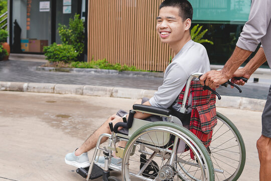 Asian man in wheelchair pushed by caregiver up ramp outdoors. Disabled son and father smiling together at public place entrance with accessible facility concept.