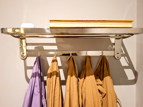 A stainless steel wall-mounted rack holding prayer garments (mukena) on hooks below, with a yellow book placed neatly on top of the shelf.