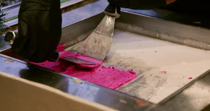 Hands in black gloves using metal spatulas to scrape and mix pink berry ice cream on a frozen metal surface, professional street food preparation, culinary process.