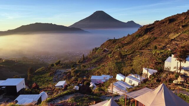 Aerial view of Sikapuk Hill and Villa Dieng featuring glamping tents and a swimming pool overlooking misty mountains and a volcano at sunrise in Indonesia.