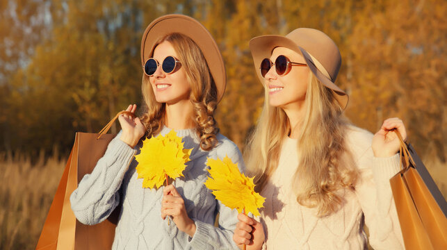 Happy two women friends with autumn yellow leaves, shopping bags, stylish girlfriends smile in park