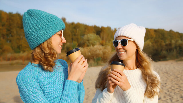 Happy young women friends talking with coffee drinks on beach, smiling girlfriends together on sea
