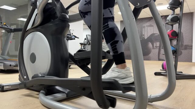 A person exercises on an elliptical machine in a gym. Other gym equipment is visible around. People can be seen working out.