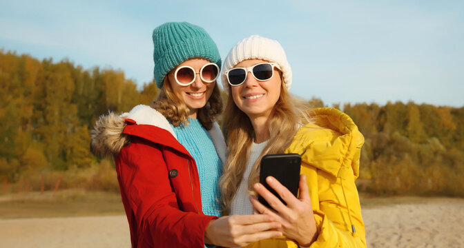 Happy young women friends with smartphone together on beach, smiling girlfriends using phone on sea