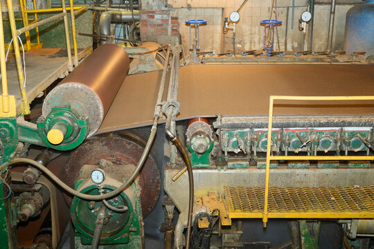 Large industrial machine rollers move wide brown sheets of cardboard through a recycling plant during daytime operations to produce new paper products