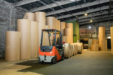 Forklift operator moves heavy industrial rolls of recycled cardboard inside a warehouse to prepare...