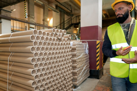 Worker wearing safety gear inspects inventory of recycled cardboard paper rolls stored in a large warehouse plant during daytime production hours