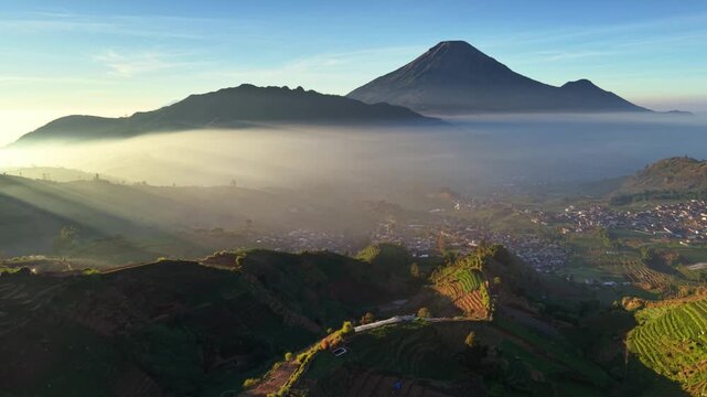 Aerial view of the misty Sikapuk Hill and terraced fields of the Dieng Plateau at sunrise in Indonesia.