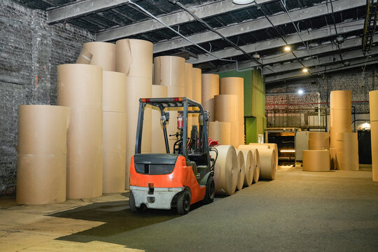 Forklift operator moves heavy industrial rolls of recycled cardboard inside a warehouse to prepare materials for paper production during the workday