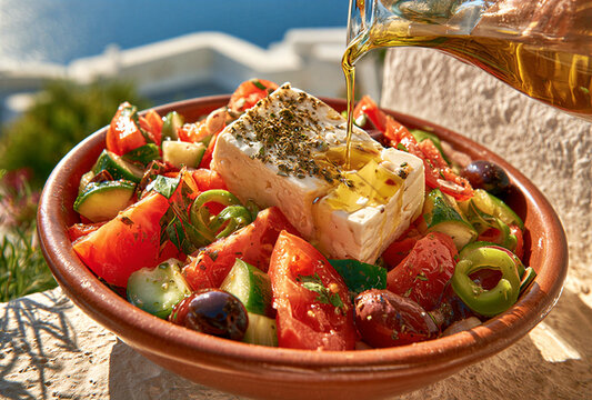 A delicious Greek salad with feta cheese and olives being drizzled with olive oil, set against a backdrop of whitewashed buildings under the sun.