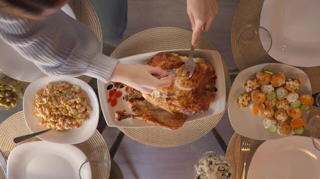 Woman carving roasted turkey during festive Christmas family dinner, serving traditional holiday meal while relatives gather around table and celebrate together at home.