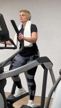 A woman uses an elliptical trainer in a gym. She wears workout clothing and a towel around her neck. The exercise takes place indoors.