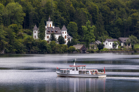 View of a white passenger boat cruising on the calm waters of Lake Hallstatt with the historic Schloss Grub castle nestled among dense green forests in Hallstatt, Upper Austria, Austria.