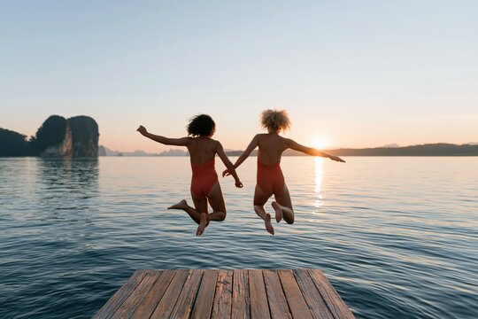 Two friends jumping off a wooden dock into a lake at sunset holding hands for summer adventure poster and travel background