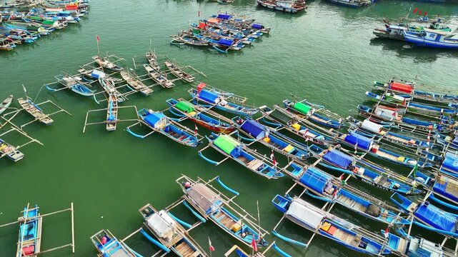 Aerial view of numerous colorful traditional fishing boats with outriggers moored closely together in the calm green waters of a harbor in Indonesia.