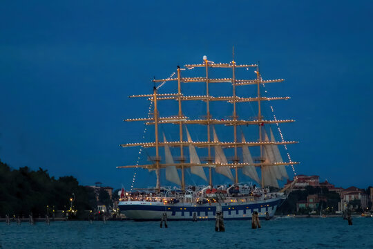 View of a large illuminated sailing ship cruising through the lagoon at dusk with the historic skyline of Venice, Venice, Veneto, Italy.
