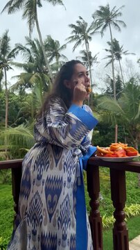 Woman in ikat pattern kimono standing on balcony eating fresh tropical papaya and fruit