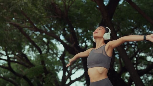 Happy young Asian woman wearing wireless headphones and sportswear stretching arms outdoors in green park, enjoying music after exercise, active healthy lifestyle, fitness, freedom and wellness