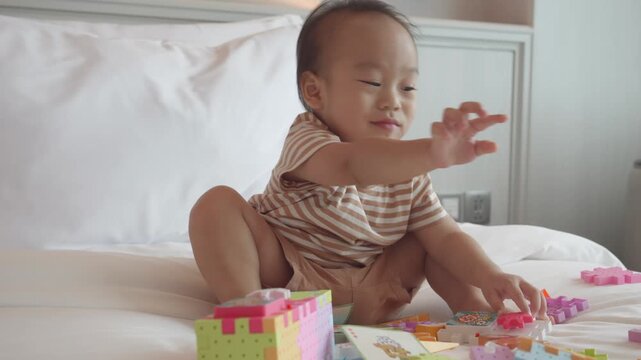 A cute baby is seen crawling on a bed covered with colorful toy blocks and a book. The soft, cozy setting creates a delightful atmosphere, highlighting the joy of childhood playtime.