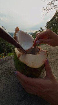 Hands Scoop Tender Coconut Flesh from an Opened Green Coconut on a Rustic Table, Framed by Forested Hills and Distant Water, Showing Fresh Tropical Food Preparation Vertical video.