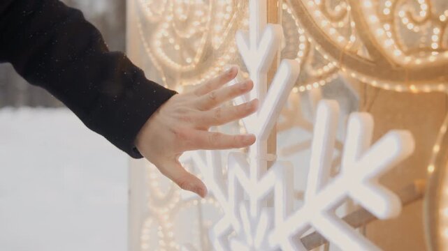 Close up of woman hand gently touching and tracing illuminated snowflake decorations on outdoor display during winter. scene features soft lighting from string lights creating warm