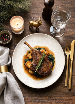 Roasted lamb chop with rosemary, vegetables, candlelight, wine glass, cutlery, and evergreen branches arranged on a rustic wooden dining table.