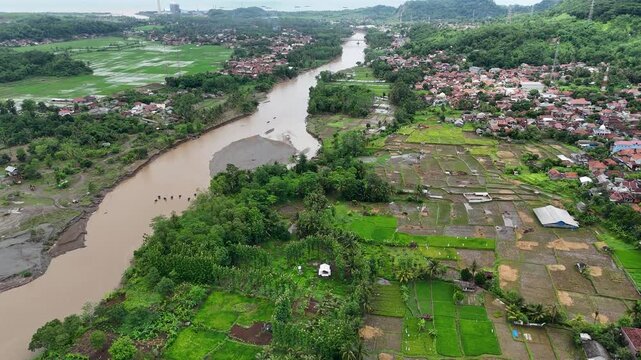 Aerial view of Lio citarik river flowing through lush green rice fields and rural village houses in Kecamatan Pelabuhanratu, Jawa Barat, Indonesia.