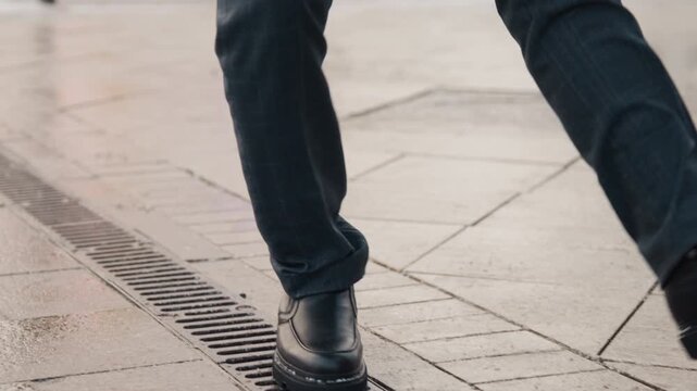 Close up of man legs and feet in black leather shoes walking on wet pavement. man wears dark pants with focus remaining solely on lower body movement against urban background