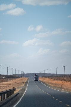 View of a vehicle driving along a straight asphalt road through the arid landscape of Amboseli National Park, lined with utility poles in Kajiado County, Kenya.