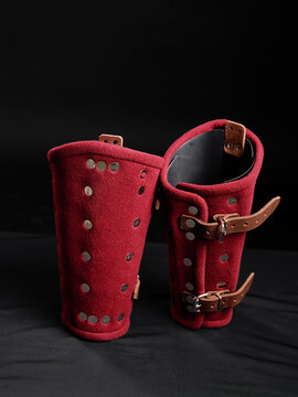 Pair of red felt greaves with metal studs and leather straps, standing upright on dark background.