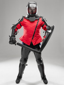 HEMA fighter posing frontally with padded sword and shield in red jacket.