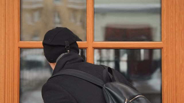 Close up of person wearing winter clothing and carrying backpack looking out through glass doors at urban scene with snow covered ground. person remains in same position throughout observing