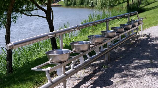 A row of stainless steel public washbasins stands beside a calm lake on a bright summer day. Concept of outdoor public sanitation, recreation infrastructure, and summer travel convenience.