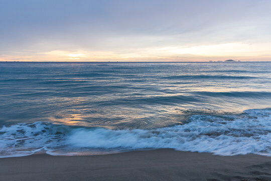 Calm ocean waves gently wash onto a sandy beach beneath a soft pastel sunrise sky, evoking serene coastal beauty, peaceful morning mood and expansive seascape with distant islands. Cam Ranh, Vietnam