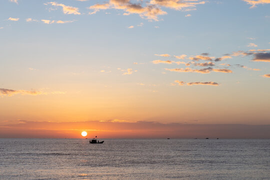 A lone fishing boat drifts on calm ocean waters as the golden sun sets beneath a pastel sky casting warm reflections and creating a peaceful reflective mood across the wide seascape. Cam Ranh, Vietnam