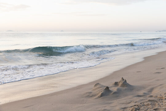 A small sandcastle near the waterline on a quiet sandy beach at sunrise, with gentle ocean waves rolling in, soft pastel sky and a calm horizon reflecting the tranquil morning mood. Cam Ranh, Vietnam