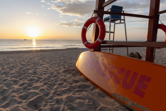 Lifeguard rescue board leaned against a wooden tower with life rings on a sandy beach at sunrise, warm golden light on calm ocean waters creating a peaceful coastal safety scene. Cam Ranh, Vietnam