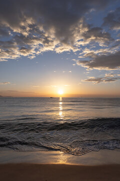A peaceful golden sunset casts warm light across a calm ocean and sandy beach, with reflective water, gentle waves and dramatic clouds creating a serene, contemplative coastal atmosphere of Cam Ranh