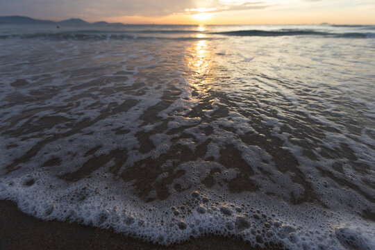 Warm golden sunset light reflects across gentle waves as sea foam laps the sandy shore, creating a peaceful coastal scene that evokes calm, solitude and the quiet beauty of nature