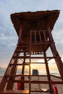 A wooden lifeguard tower overlooks a quiet sandy beach at sunrise, the sun glinting on the ocean horizon and casting warm golden light over waves, sand and a peaceful coastal scene. Vertical photo
