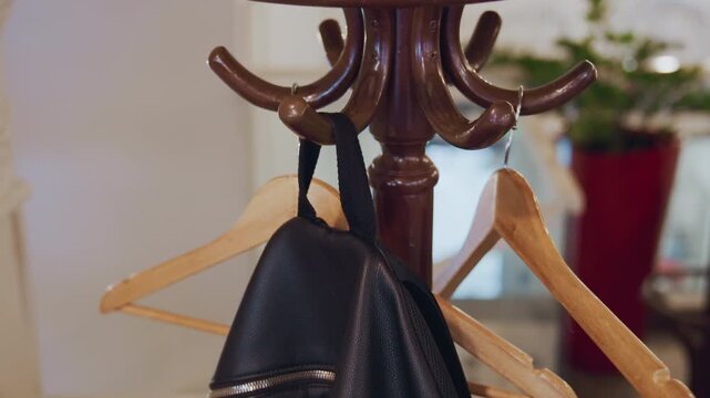 Close up of person hanging black leather backpack on wooden hangers attached to brown coat rack in indoor setting. Hand with dark sleeve reaches for hook places bag securely among other