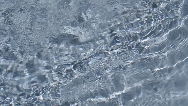 Clear rippling water surface creating bright light refractions over a smooth stony bottom. Concept of refreshing summer relaxation, pure liquid textures, and natural tranquility.