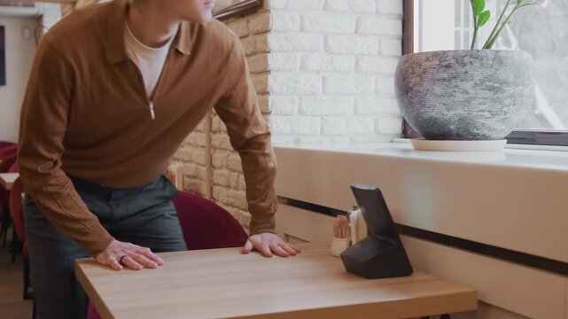 Man in brown sweater moves to sit at empty table with red chairs and white brick walls. Man adjusts chair places his hands on wooden tabletop near condiment holders like salt shakers