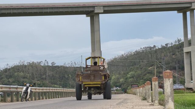 Self-made truck driving on a rural road with a modern concrete highway overpass bridge in the Vietnamese countryside