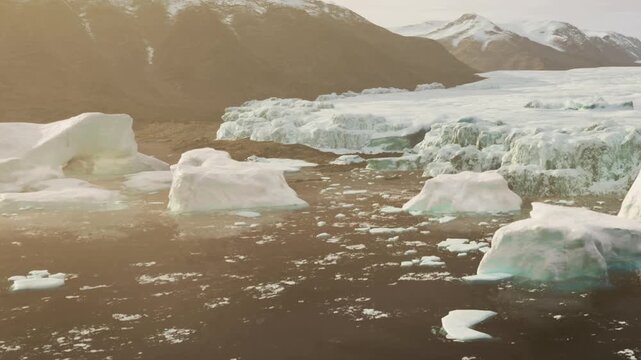 Large icebergs float in a body of water surrounded by mountains. The sun shines above, creating a bright scene with reflections on the ice.