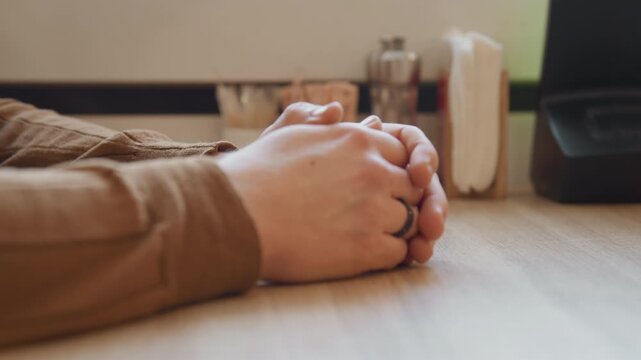 Close up of man resting his clasped hands on edge of wooden table in indoor workspace. scene includes soft lighting and visible objects like books containers with utensils indicating calm